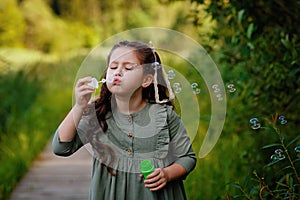 A girl with dark hair on a background of green foliage launches soap bubbles. Summer outdoor activities. Blow a soap bubble