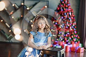 Girl with dark hair standing on a box with gifts. Christmas tree in the background. smiles