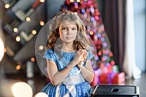 Girl with dark hair standing on a box with gifts. Christmas tree in the background. smiles