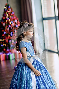 Girl with dark hair standing on a box with gifts. Christmas tree in the background. smiles