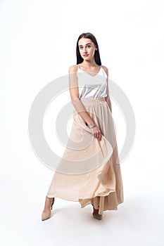 A girl with dark hair in a white top and a long beige skirt poses on a white background