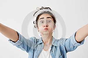 Girl with dark and wavy hair in bun stretching arms to camera, isolated against white background, pouting cheeks. Trendy