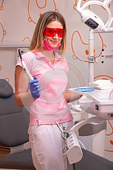 A girl dentist wearing red glasses and a photo lamp in her hands, smiling in the dental office.