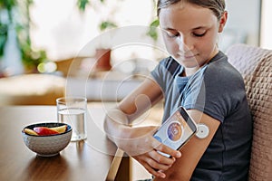 Girl with diabetes checking blood glucose level at home using continuous glucose monitor. Schoolgirl connecting CGM to