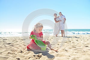 Girl digging sand on beach with parents in background