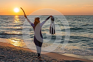 A girl diver on beach meets dawn raising hands up. Woman wearing wetsuit and holding diving equipment fins and snorkel