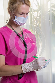 A girl doctor in a mask and gloves writes down the readings of the patient's temperature and pressure in a notebook.