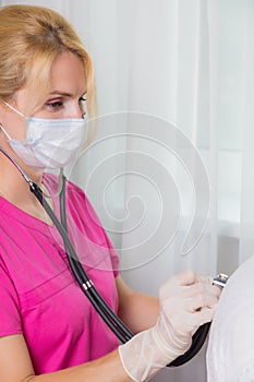 A girl doctor in a mask listens with a stethoscope to the back of an adult woman.