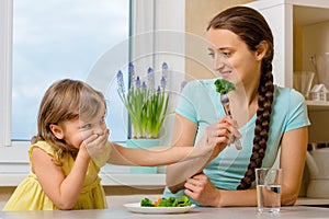 Girl doesn`t want eat healthy vegetables. Kid looks with disgust at broccoly. Mother convinces her daughter to eat food