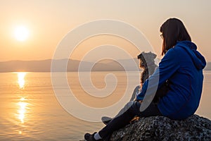 Girl with dog looking sunset on sea, lake. Woman sitting on rocks. Lonely person. Girl alone outside. Vacation on sea