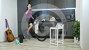 Girl doing sports exercises on cycling trainer and drinking water or isotonic from bottle during training. Woman cycling indoor on