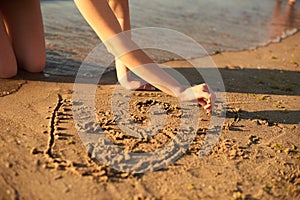 The girl is drawing a heart on the sand by the sea in the evening with fingers