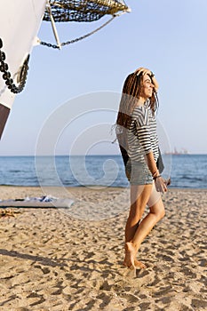 A girl with a dreadlocked hairstyle poses on the beach near a ship in summer, the sea and sand on a bright sunny day, dressed in a