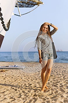 a girl with a dreadlocked hairstyle poses on the beach near a ship in summer, the sea and sand on a bright sunny day, dressed in a