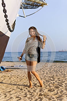 a girl with a dreadlocked hairstyle poses on the beach near a ship in summer, the sea and sand on a bright sunny day, dressed in a