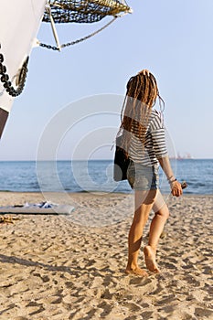 a girl with a dreadlocked hairstyle poses on the beach near a ship in summer, the sea and sand on a bright sunny day, dressed in a