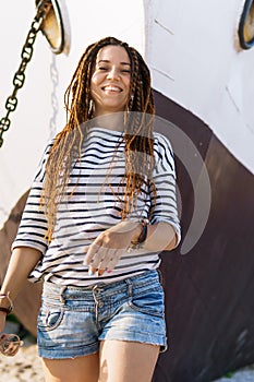 A girl with a dreadlocked hairstyle poses on the beach near a ship in summer, a sunny day, dressed in a T-shirt and denim shorts
