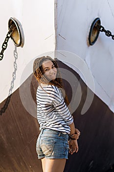 a girl with a dreadlocked hairstyle poses on the beach near a ship in summer, a sunny day, dressed in a T-shirt and denim shorts