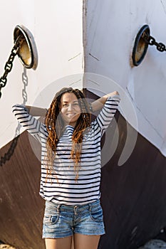 a girl with a dreadlocked hairstyle poses on the beach near a ship in summer, a sunny day, dressed in a T-shirt and denim shorts