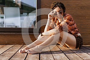 a girl with a dreadlocked hairstyle poses on the beach in summer against a red brown wooden wall, bright sunlight, dressed in a