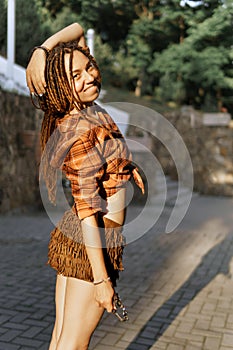 a girl with a dreadlocked hairstyle poses in summer outdoor, bright sunlight, dressed in a plaid shirt and shorts