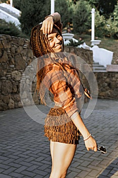 a girl with a dreadlocked hairstyle poses in summer outdoor, bright sunlight, dressed in a plaid shirt and shorts