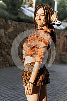 a girl with a dreadlocked hairstyle poses in summer outdoor, bright sunlight, dressed in a plaid shirt and shorts