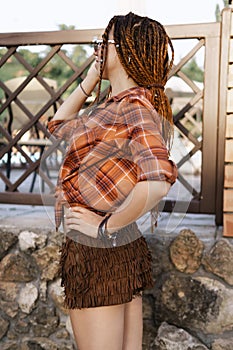 a girl with a dreadlocked hairstyle poses in summer outdoor, bright sunlight, dressed in a plaid shirt and shorts