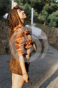 a girl with a dreadlocked hairstyle poses in summer outdoor, bright sunlight, dressed in a plaid shirt and shorts