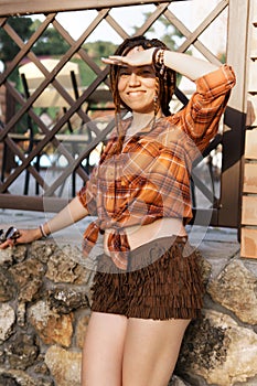 a girl with a dreadlocked hairstyle poses in summer outdoor, bright sunlight, dressed in a plaid shirt and shorts