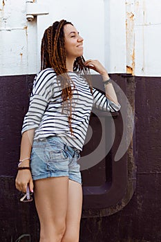 a girl with a dreadlocked hairstyle poses in the summer outdoors, dressed in a T-shirt and denim shorts