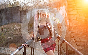 A girl with dreadlocks in a leather jacket and a short skirt stands against the background of an old stone wall in the rays of a b