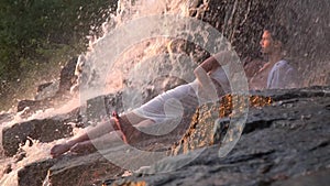 A girl in a dress kisses a guy in the neck and falls lower while they lie on the rocks under a waterfall during the