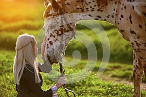 Girl in dress strokes a horse in summer background with green grass