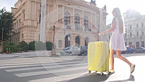 The girl is dressed in a dress and a white hat, walks down the street and pushes a suitcase.