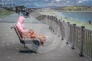 Girl dressed in jacket and wrapped in blanket sits with a glass of coffee on the bench, shore of a cold gloomy sea.