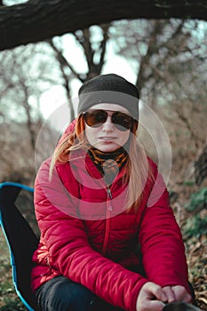 The girl is dressed in a red down jacket holding metal cup. Sunglasses and hat. Sits in a folding camping chair. Light weight and