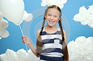 Girl dressed in striped dress posing on a blue background with cotton clouds, white air balloon, the concept of summer and happine