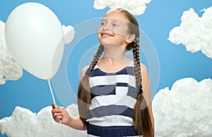 Girl dressed in striped dress posing on a blue background with cotton clouds, white air balloon, the concept of summer and happine