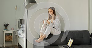 Girl drinking coffee or tea on sofa. Woman working, learns and using laptop computer in the room. Freelance. Writing