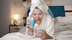 Girl drinking coffee or tea and use phone. Attractive woman in bedroom in white bathrobe lies in bed drinks hot coffee