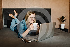 Girl drinking coffee and watching movie in home. Woman working, learns and using laptop computer in the bedroom. Freelancer.