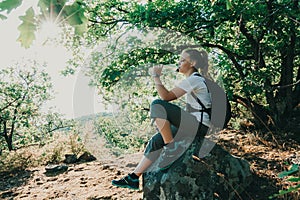 Girl drinking mineral water sitting on stone in forest. Hiker resting with aqua bottle during hike. Thirst quench beverage