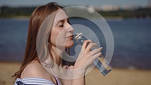 A girl drinks water from a plastic bottle while sitting on a sandy beach near a river.