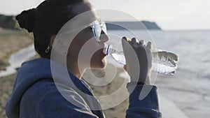 The girl drinks water from a transparent bottle after jogging on the seashore
