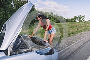 Girl driver near the open hood of a broken car. Broken car on the roadside with an open hood, trunk. Woman repairing the