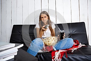 Girl eating chips, drinking soda, watching tv, sitting on sofa.