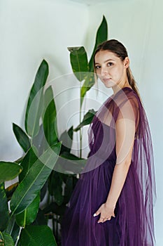 Girl in elegant long dress with a curious questioning look stands at the green exotic leaves