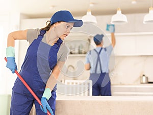 Girl employee of cleaning company in blue jumpsuit cleans and washes floor in kitchen