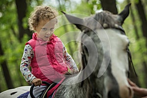 Girl enjoying horseback riding in the woods, young pretty girl with blond curly hair on a horse with backlit leaves